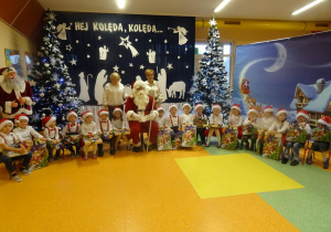 Grupa dzieci z prezentami w ręku, Mikołajem i panią dyrektor Maria Królikowska i panią Agnieszka Piekarska na tle świątecznej dekoracji.